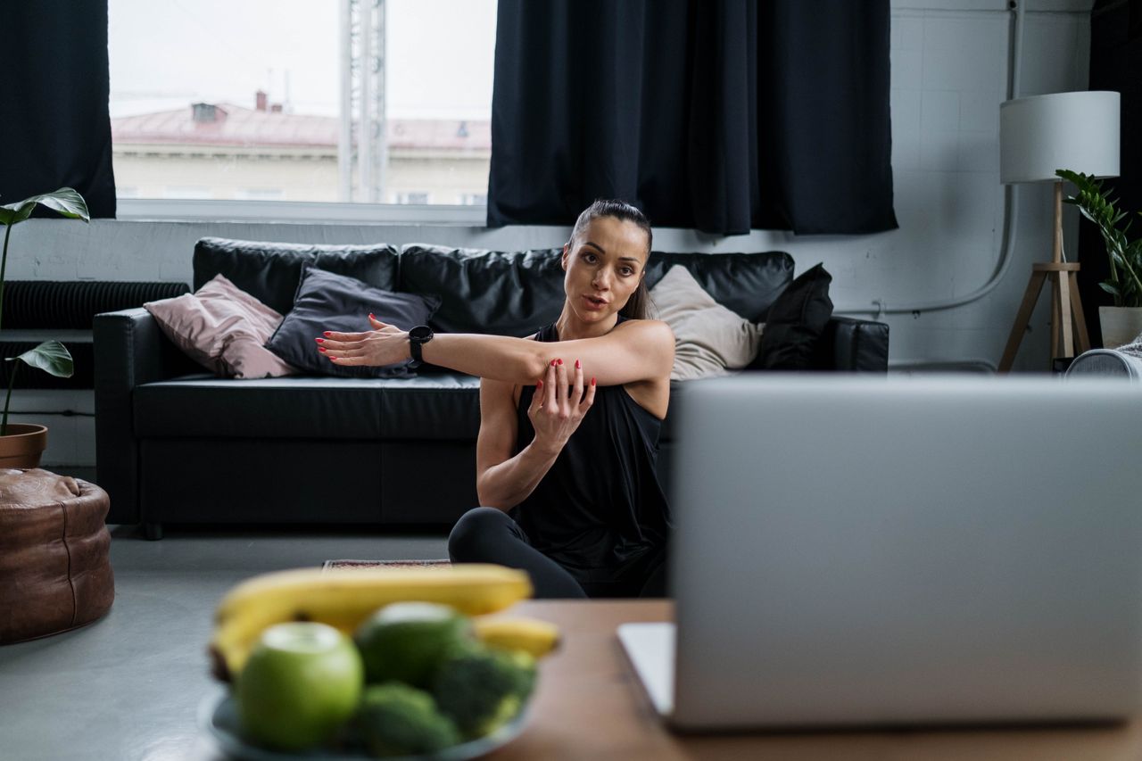 Home Woman practicing yoga at home with a laptop, stretching arms in a cozy living room setting.